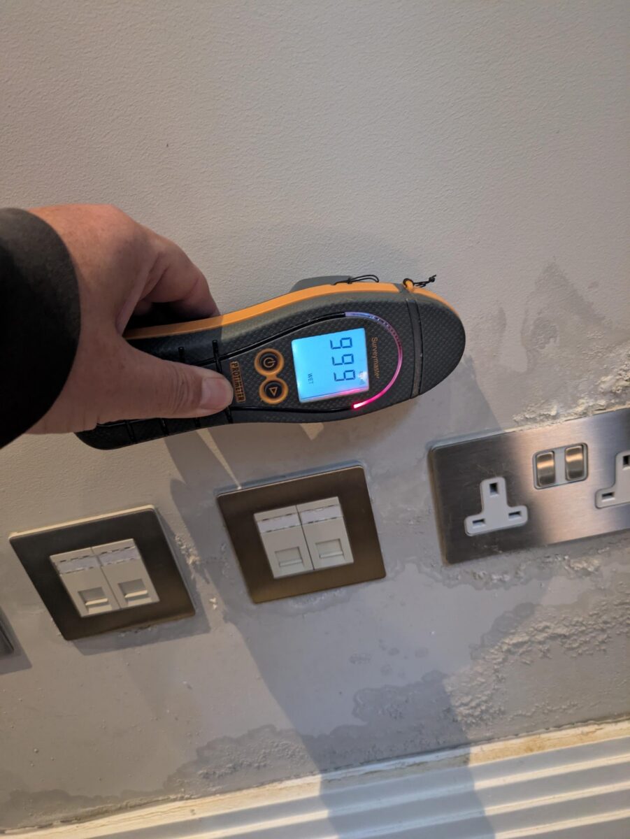 Moisture meter showing the maximum 999 reading on a water-damaged lounge wall